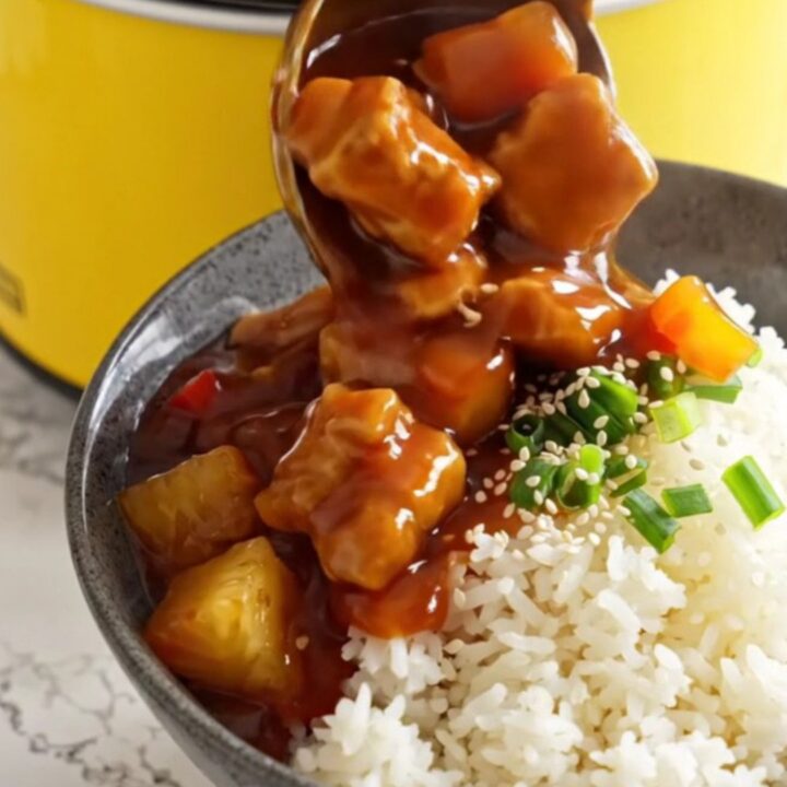 A ceramic slow cooker filled with sweet and sour pork, pineapple, and bell peppers in a glossy red sauce.