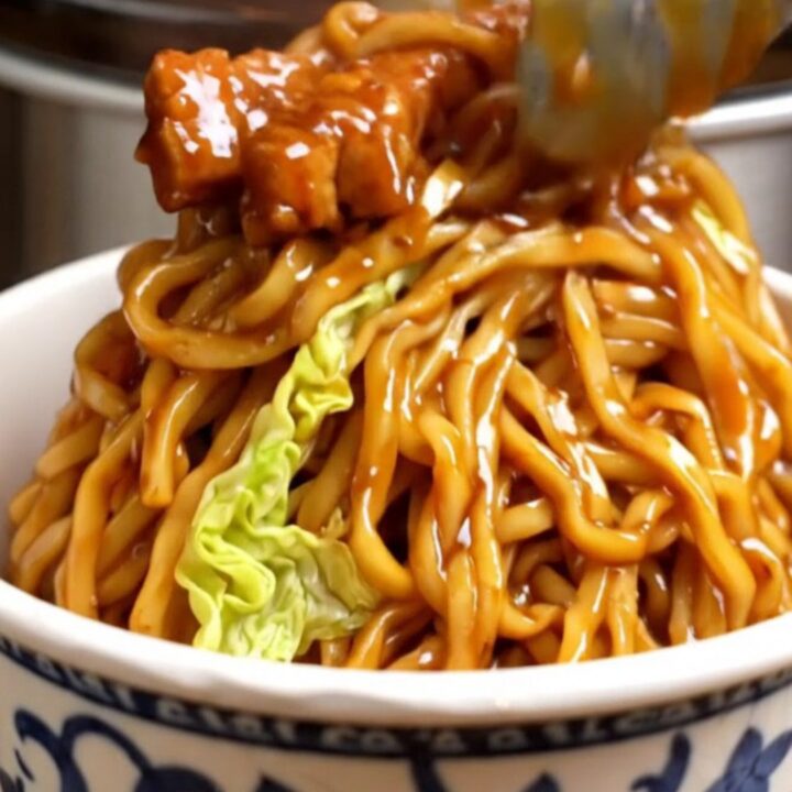 A close-up of glossy Char Siu pork lo mein noodles in a takeout bowl with mushrooms and carrots.
