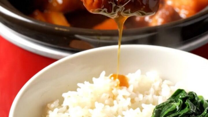 A close-up view of Shanghai red-braised pork belly in a red ceramic crockpot, showing mahogany-glazed cubes.