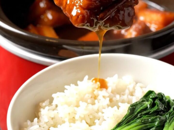 A close-up view of Shanghai red-braised pork belly in a red ceramic crockpot, showing mahogany-glazed cubes.
