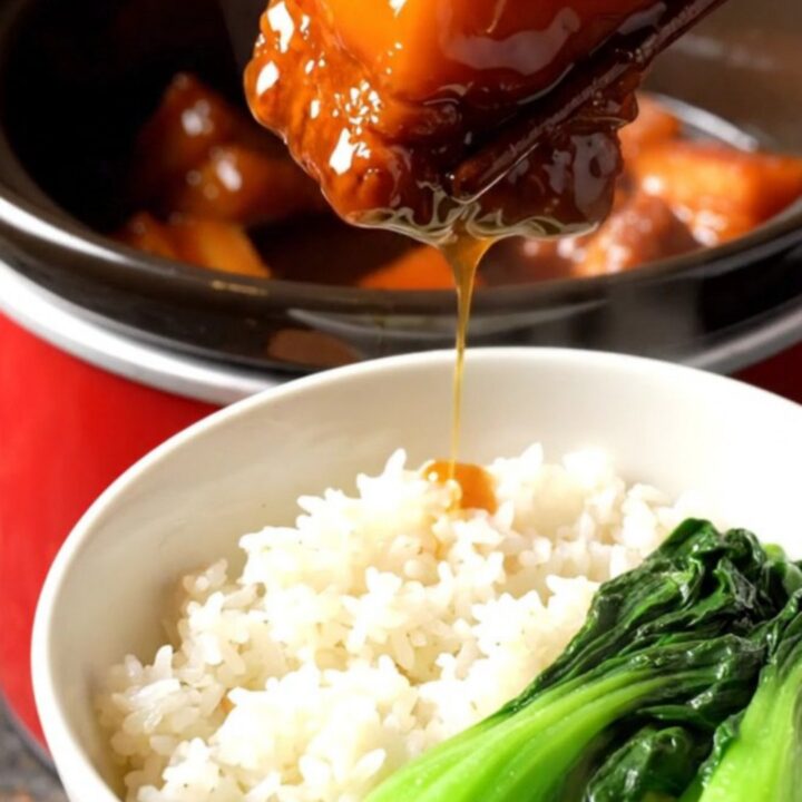 A close-up view of Shanghai red-braised pork belly in a red ceramic crockpot, showing mahogany-glazed cubes.