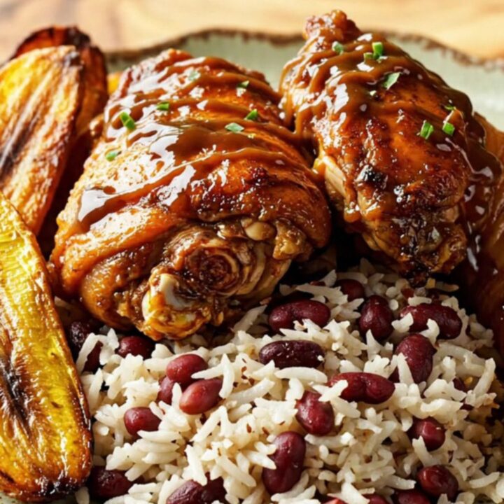 A close-up of glazed Jamaican jerk chicken thighs served on a plate with coconut rice and plantains.