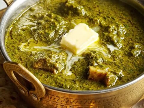 A close-up of a brass bowl filled with creamy Saag Gosht, showing tender lamb chunks and golden paneer in a vibrant green