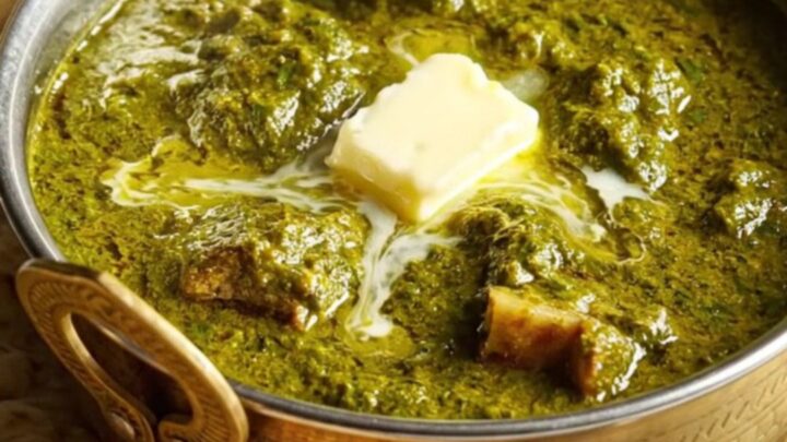 A close-up of a brass bowl filled with creamy Saag Gosht, showing tender lamb chunks and golden paneer in a vibrant green