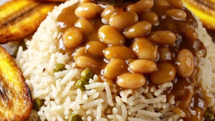 A close-up of tender Jamaican brown stew oxtail with butter beans served over rice in a dark ceramic bowl.