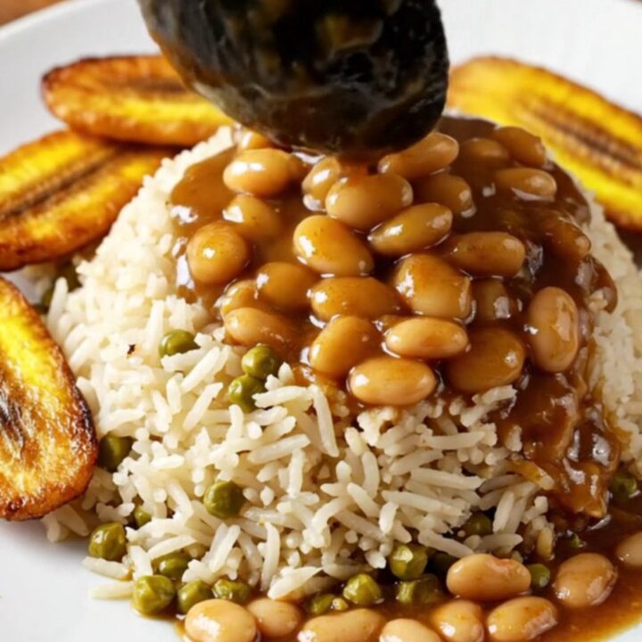 A close-up of tender Jamaican brown stew oxtail with butter beans served over rice in a dark ceramic bowl.