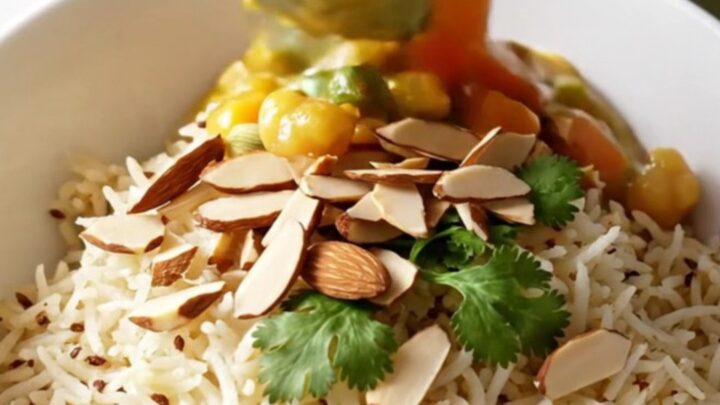A close-up of a bowl of creamy Vegetable Korma served over basmati rice with almonds and cilantro.
