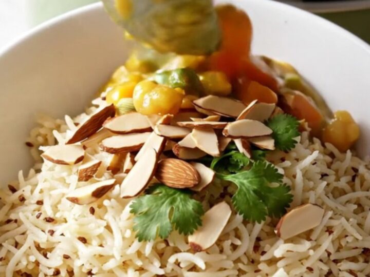 A close-up of a bowl of creamy Vegetable Korma served over basmati rice with almonds and cilantro.