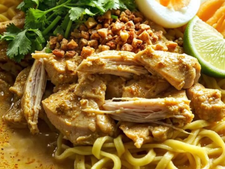 A steaming bowl of golden Burmese coconut chicken curry with egg noodles and fresh herbs.