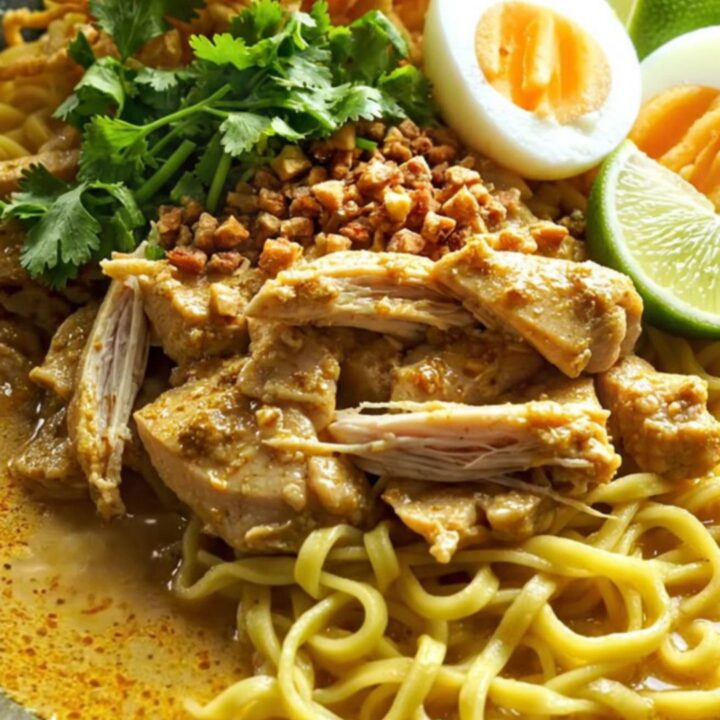 A steaming bowl of golden Burmese coconut chicken curry with egg noodles and fresh herbs.