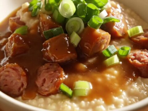 Close up of a ceramic crockpot filled with creamy cheddar grits topped with smoky andouille sausage and gravy.