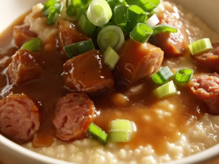 Close up of a ceramic crockpot filled with creamy cheddar grits topped with smoky andouille sausage and gravy.