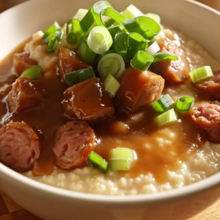Close up of a ceramic crockpot filled with creamy cheddar grits topped with smoky andouille sausage and gravy.