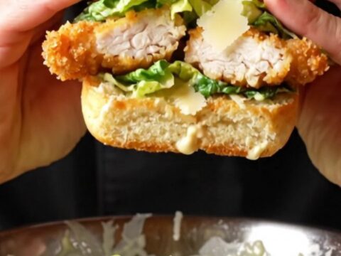 Crispy chicken Caesar sandwich on a wooden board, cut in half showing layers of chicken, romaine, and parmesan.