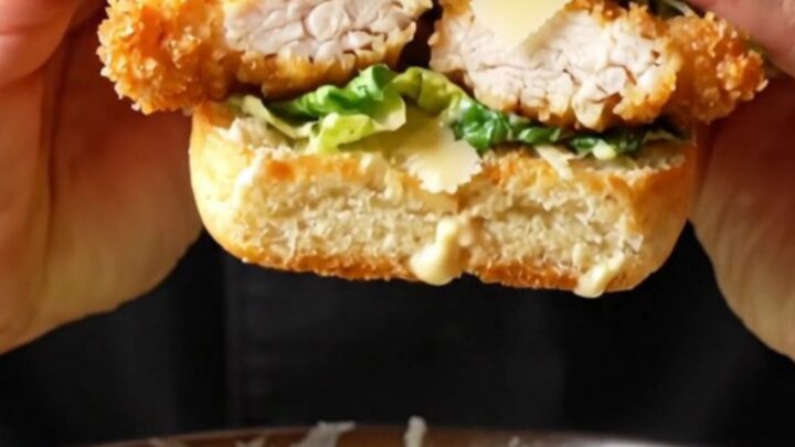 Crispy chicken Caesar sandwich on a wooden board, cut in half showing layers of chicken, romaine, and parmesan.