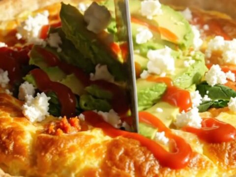 A golden puffed tortilla-crust quiche topped with avocado slices, cotija, and hot sauce in a ceramic baking dish