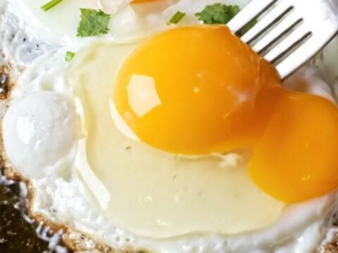 Fork breaking a runny fried egg over red chile beef enchiladas, golden yolk cascading into brick-red sauce and melted cheddar