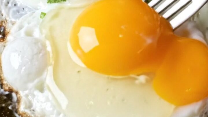 Fork breaking a runny fried egg over red chile beef enchiladas, golden yolk cascading into brick-red sauce and melted cheddar