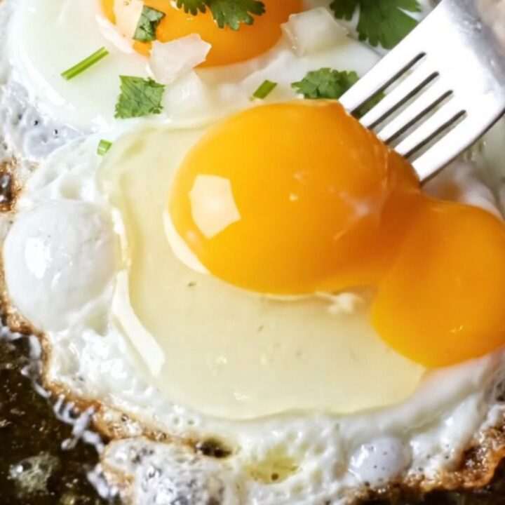 Fork breaking a runny fried egg over red chile beef enchiladas, golden yolk cascading into brick-red sauce and melted cheddar