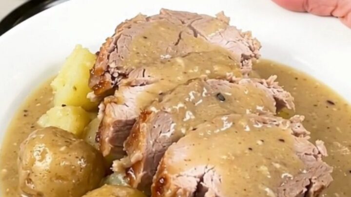 Slices of crockpot pork loin with soft baby potatoes under thick creamy bacon gravy on a white plate