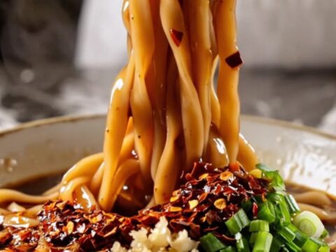 Wide glossy Biang Biang belt noodles coated in fiery red chili oil with bok choy, lifted by chopsticks with steam rising