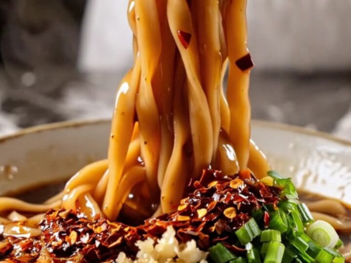 Wide glossy Biang Biang belt noodles coated in fiery red chili oil with bok choy, lifted by chopsticks with steam rising