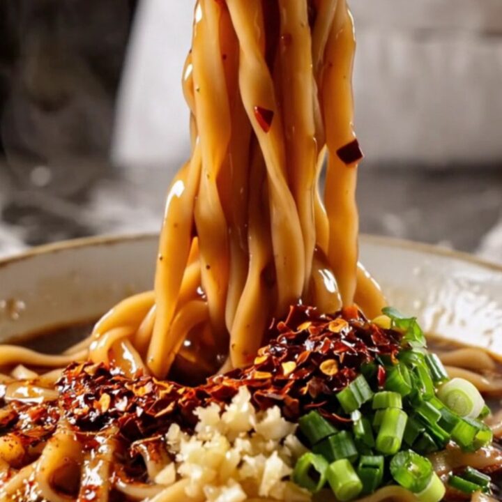 Wide glossy Biang Biang belt noodles coated in fiery red chili oil with bok choy, lifted by chopsticks with steam rising