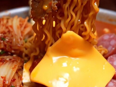 Shallow pot of Korean army stew ramyun with brick-red noodles draped in melted cheddar, pork belly and kimchi