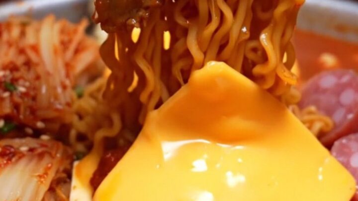 Shallow pot of Korean army stew ramyun with brick-red noodles draped in melted cheddar, pork belly and kimchi