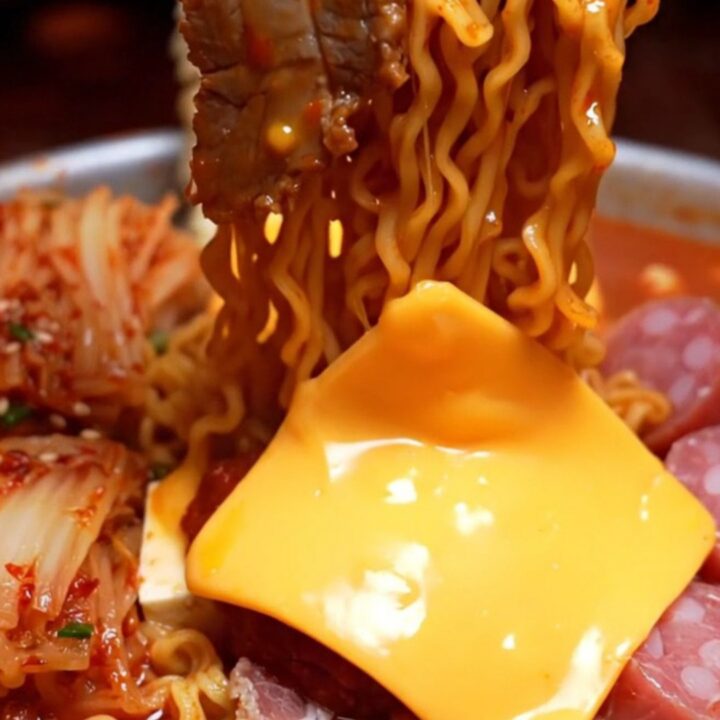 Shallow pot of Korean army stew ramyun with brick-red noodles draped in melted cheddar, pork belly and kimchi