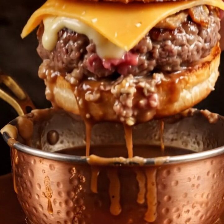 French Dip smash burger with melted Gruyère and crispy onions being dunked into a steaming bowl of rosemary au jus