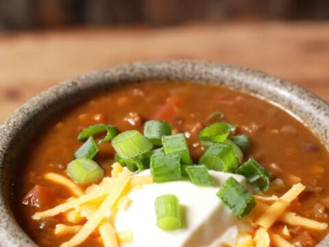 Ladle lifting espresso-dark crockpot venison chili with black beans and a melted aged cheddar pull from a slow cooker