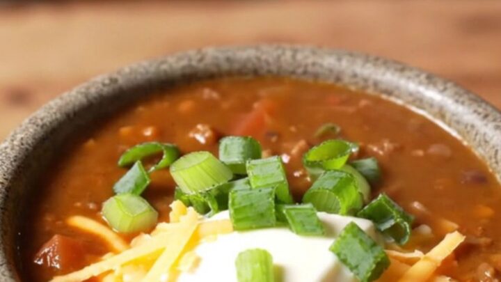Ladle lifting espresso-dark crockpot venison chili with black beans and a melted aged cheddar pull from a slow cooker
