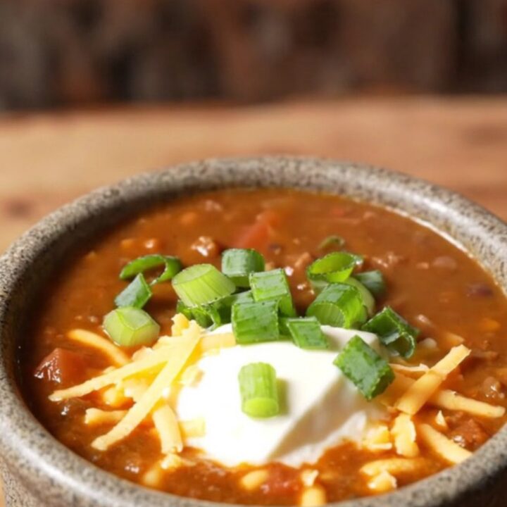 Ladle lifting espresso-dark crockpot venison chili with black beans and a melted aged cheddar pull from a slow cooker
