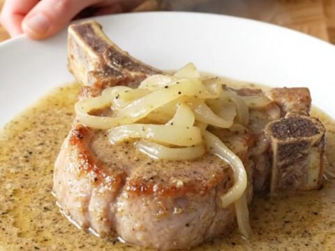 Bone-in pork chop on a white plate under soft onions and thick creamy peppered country gravy, steam rising
