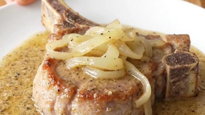 Bone-in pork chop on a white plate under soft onions and thick creamy peppered country gravy, steam rising