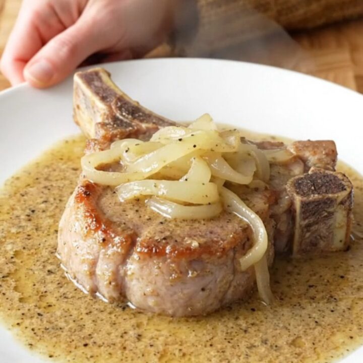 Bone-in pork chop on a white plate under soft onions and thick creamy peppered country gravy, steam rising