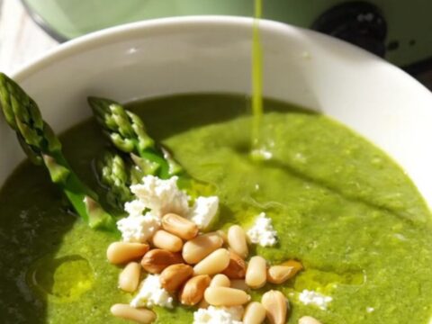 Emerald asparagus bisque in a white ceramic bowl topped with goat cheese crumbles, pine nuts, and asparagus tips