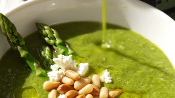 Emerald asparagus bisque in a white ceramic bowl topped with goat cheese crumbles, pine nuts, and asparagus tips