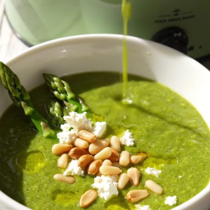 Emerald asparagus bisque in a white ceramic bowl topped with goat cheese crumbles, pine nuts, and asparagus tips