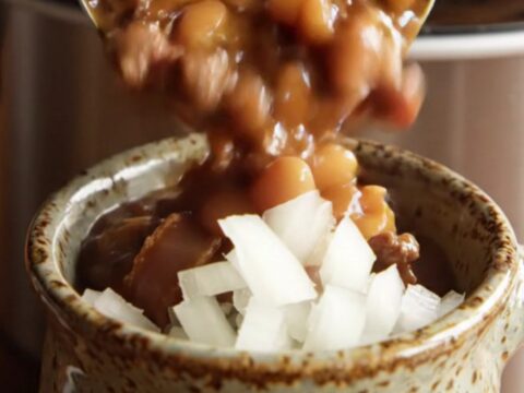 Ladle lifting hearty cowboy calico beans with ground beef, bacon and three beans from a bubbling slow cooker
