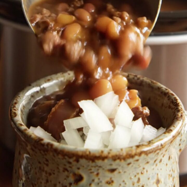 Ladle lifting hearty cowboy calico beans with ground beef, bacon and three beans from a bubbling slow cooker
