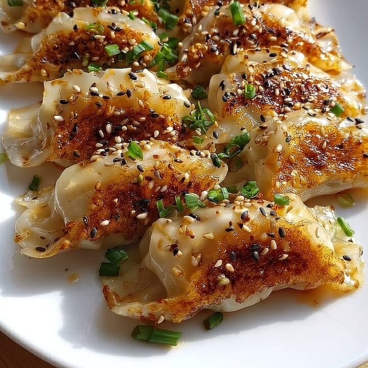 Gyozas poêlés dorés en rosace sur une assiette ovale blanche, nappage brillant, graines de sésame et cives