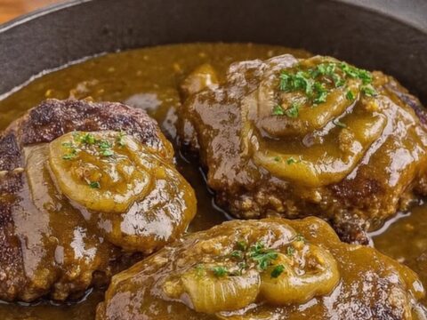 Three smothered hamburger steaks glistening with onion gravy in a cast-iron skillet