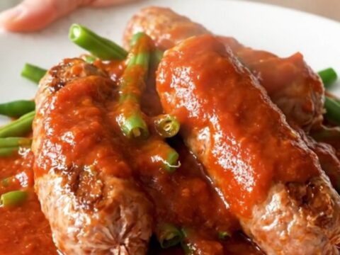 Whole Italian sausage links and green beans in thick red marinara sauce on a white plate, steam rising