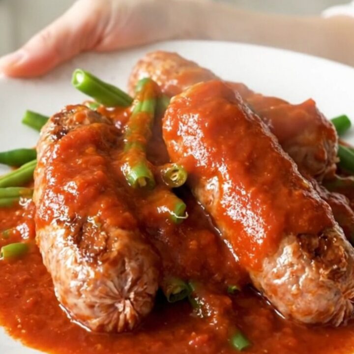 Whole Italian sausage links and green beans in thick red marinara sauce on a white plate, steam rising