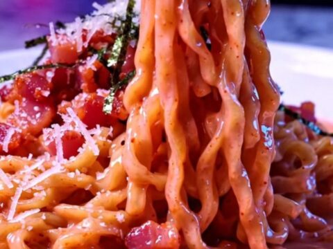 Glossy pink spicy ramen carbonara noodles twirled in a bowl with crisp bacon, Parmesan and nori strips