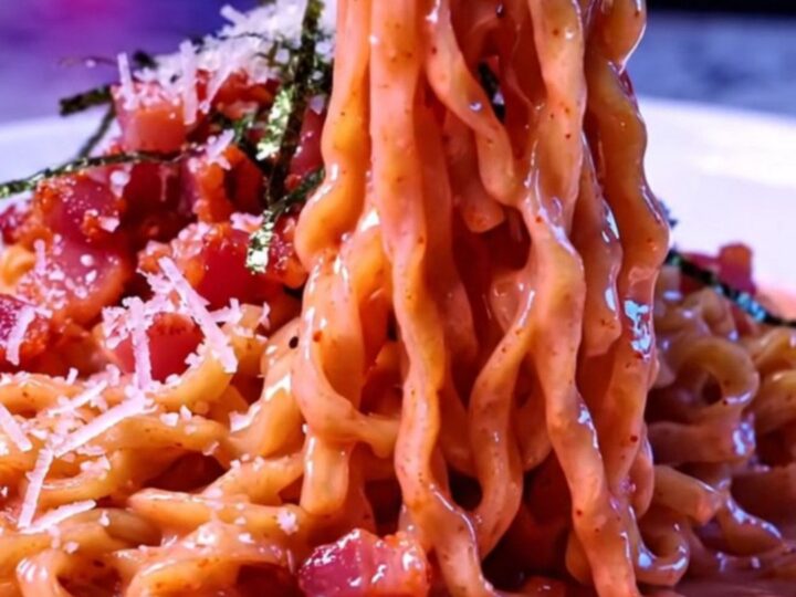 Glossy pink spicy ramen carbonara noodles twirled in a bowl with crisp bacon, Parmesan and nori strips