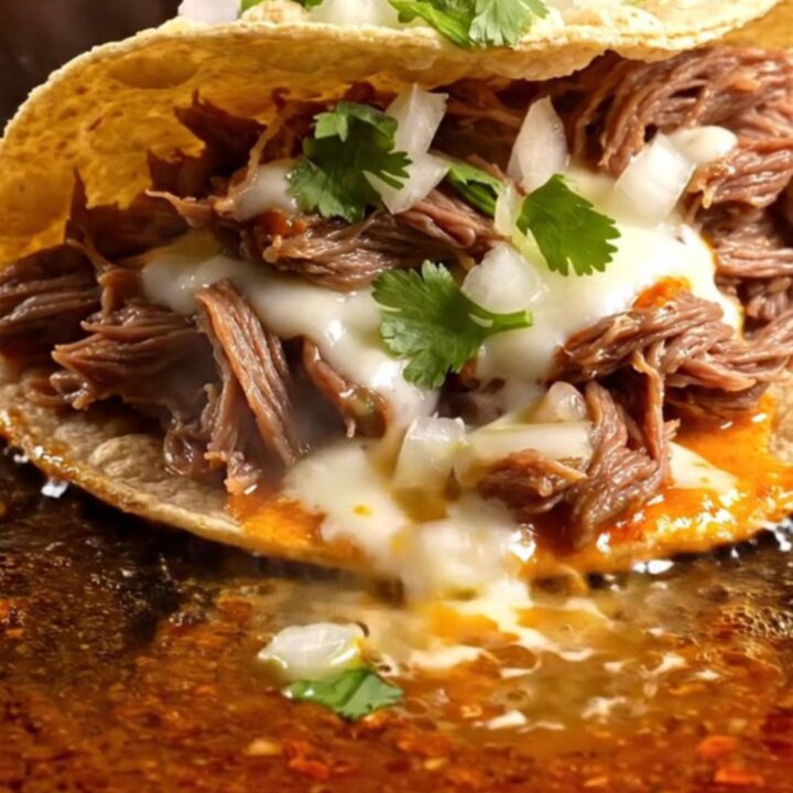 Two crispy quesabirria tacos with molten Oaxaca cheese being dipped into a bowl of glossy red beef consomé