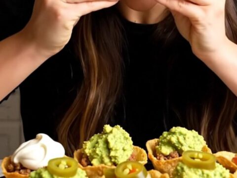 Golden baked mini taco cups topped with sour cream, guacamole and pickled jalapeño slices, one lifted with a long cheese pull